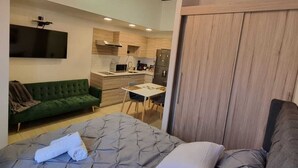 A modern bedroom with a large bed, a green sofa, a dining area, and a kitchen in the background.