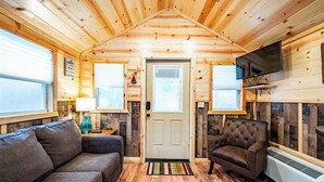 A cozy cabin interior with a wooden ceiling, a sofa, a chair, a television, and a door. - Willow: Sleeps 3, Tiny Cabin, Pets, Near Lake/Park (Broken Bow)