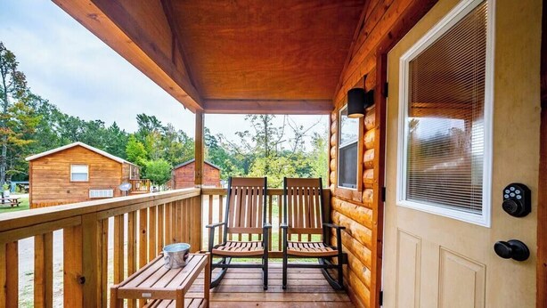 A wooden porch with two chairs and a table, a door with a window, and a view of the surrounding forest. - Willow: Sleeps 3, Tiny Cabin, Pets, Near Lake/Park (Broken Bow)