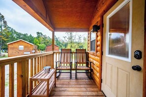 Une véranda en bois avec deux chaises et une table, une porte avec une fenêtre, et une vue sur la forêt environnante.