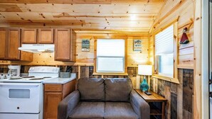 A cozy cabin-style kitchen with wooden walls, a sofa, and a window with blinds. - Willow: Sleeps 3, Tiny Cabin, Pets, Near Lake/Park (Broken Bow)