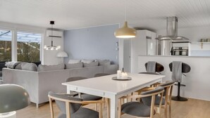 Eine moderne Küche mit weißen Schränken, eine Essecke mit weißem Tisch und grau gepolsterten Stühlen sowie ein graues Sofa im Wohnbereich.