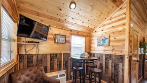 Um quarto com painéis de madeira, uma televisão, um letreiro com a palavra 'vibes', uma mesa com dois bancos e uma poltrona marrom grande.