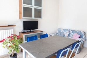 Una stanza con un tavolo da pranzo, sedie blu, una televisione su un mobile in legno, un divano con rivestimento floreale e una pianta in vaso con fiori rossi.