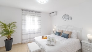 Un dormitorio con una cama, mesitas de noche, una planta en maceta, una ventana con cortinas y un aire acondicionado de pared.