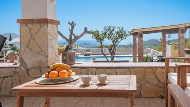 Una mesa de madera con frutero y tazas en un patio con vista a una piscina y al océano.