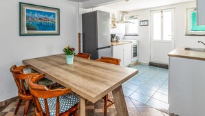 Un comedor con mesa y sillas de madera, un refrigerador, una pequeña cocina con lavamanos y un cuadro enmarcado en la pared.