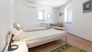 Una habitación pequeña y bien iluminada con dos camas de madera, un escritorio y una silla. Hay ventanas que dejan entrar luz natural y un cuadro en la pared.