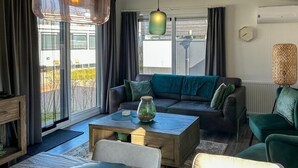 A modern living room with a sofa, coffee table, and chairs. There is a large window with curtains, a hanging light fixture, and a radiator. - Awesome home in Scherpenisse with WiFi (Scherpenisse)