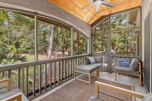 A covered outdoor seating area with a ceiling fan, wooden furniture, and a view of a landscaped garden.
