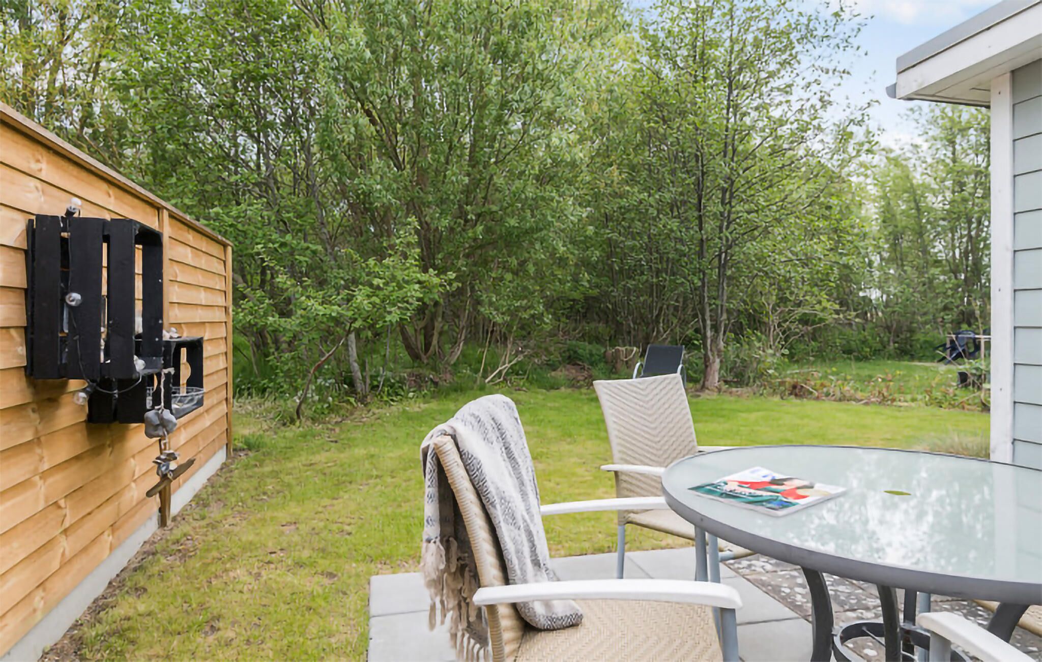Une terrasse avec une table ronde en verre et des chaises, un mur en bois avec une grille noire, et une zone herbeuse avec des arbres.