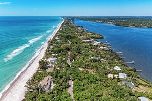 An aerial view of a coastal property with a long beachfront, dense greenery, and a dock extending into the water.