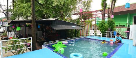 A swimming pool with a blue tiled deck, surrounded by trees and a covered area with chairs.