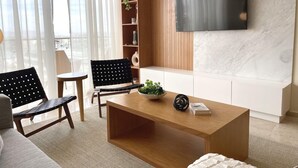 En moderne stue med et træsofabord, to sorte lænestole, en hvid sofa, et tv monteret på en marmorvæg og en reol med planter.