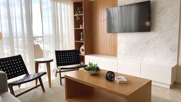 A modern living room with a wooden coffee table, two black chairs, a white sofa, a TV mounted on a marble wall, and a bookshelf with plants.