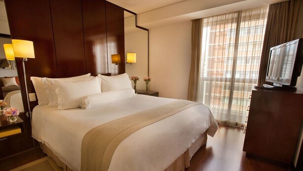 A large bed with white linens, a wooden headboard, and a beige throw blanket. There is a nightstand with a lamp and a vase of flowers. A television is on a wooden cabinet to the right. - Etoile Hotels - George V - Itaim Bibi (São Paulo)