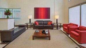 A spacious living area with a brown sofa, red armchairs, and a coffee table. There are two side tables with lamps, a large abstract painting on the wall, and a window providing natural light. - Etoile Hotels - George V - Itaim Bibi (São Paulo)