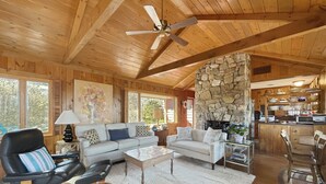 Un vaste espace de vie agrémenté d’une cheminée en pierre, d’un plafond en bois et d’un coin salon confortable comprenant un canapé et des fauteuils.