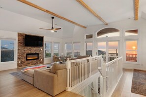 A spacious living room with a fireplace, a ceiling fan, and large windows offering views of the sunset.