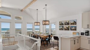 Una cocina espaciosa con comedor y vistas al océano, con vigas de madera, lámparas colgantes y obras de arte costeras.