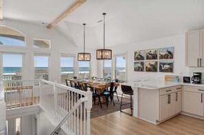 A spacious kitchen with a dining area overlooking the ocean, featuring wooden beams, pendant lights, and coastal artwork.