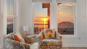 Un salón con dos sillones estampados, una mesita de noche con lámpara y ventanas amplias con vistas al atardecer.