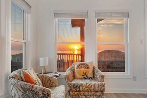 A living room with two patterned armchairs, a side table with a lamp, and large windows with a sunset view.