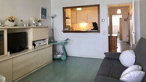 Una sala de estar con televisión, sofá, una mesa de centro y una cocina visible a través de un acceso abierto.