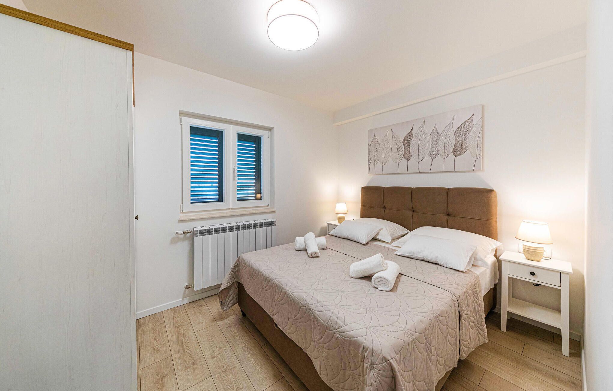 A bedroom with a bed, two bedside tables, a radiator, a window with blinds, and a wall art piece.