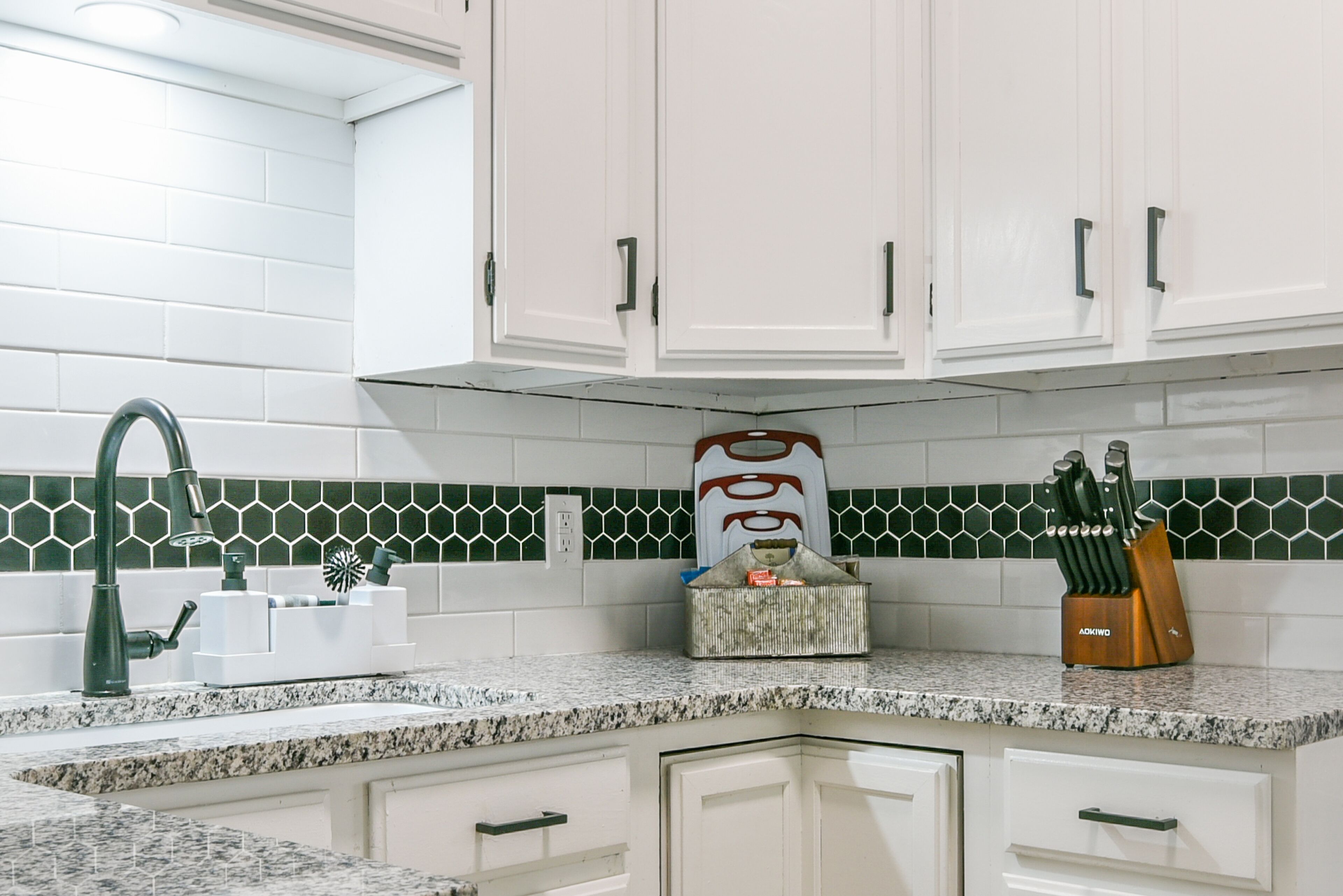 Una cocina con armarios blancos, encimera de granito, salpicadero de azulejos verdes y blancos, un bloque de cuchillos y un dispensador de jabón para platos.