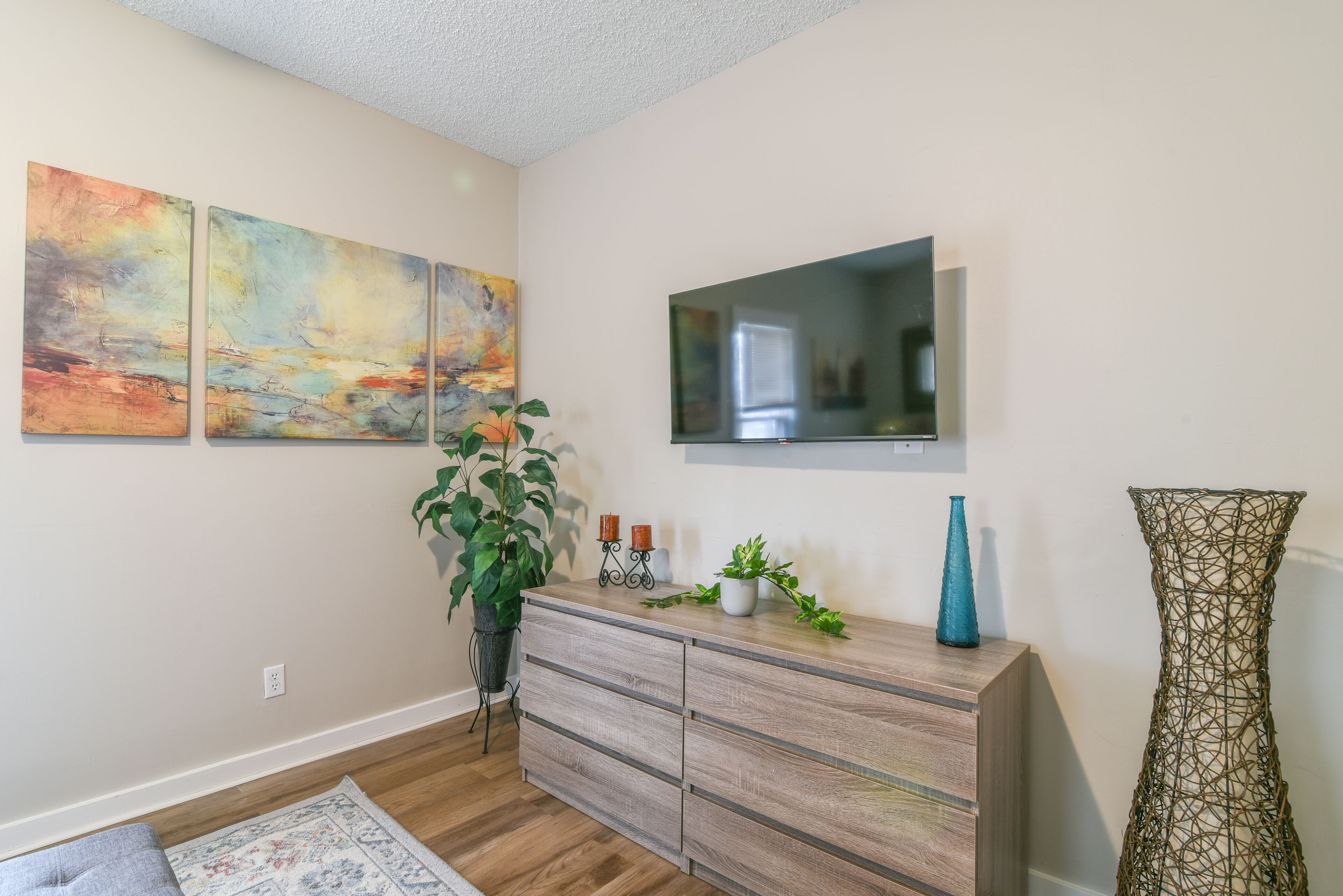 Un salón con suelo de madera, un televisor de pantalla plana montado en la pared, un mueble de madera con cajones y un cuadro abstracto de gran tamaño.