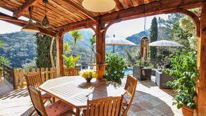 Una pérgola de madera al aire libre con una mesa y sillas, rodeada de plantas en macetas y sombrillas, con vistas a un paisaje montañoso.