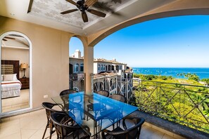 A balcony with a glass table and chairs, overlooking a coastal view with buildings and boats. - Oceanview Penthouse in Heart of Tamarindo - Perfect Location & Comfort (Tamarindo)