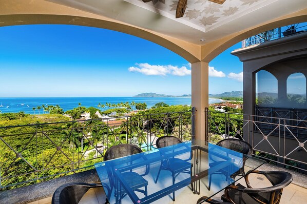 A balcony with a glass table and chairs, offering a view of the ocean and greenery. - Oceanview Penthouse in Heart of Tamarindo - Perfect Location & Comfort (Tamarindo)