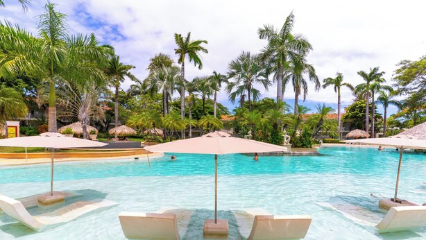 A pool area with lounge chairs and umbrellas, surrounded by palm trees and a clear sky. - Oceanview Penthouse in Heart of Tamarindo - Perfect Location & Comfort (Tamarindo)