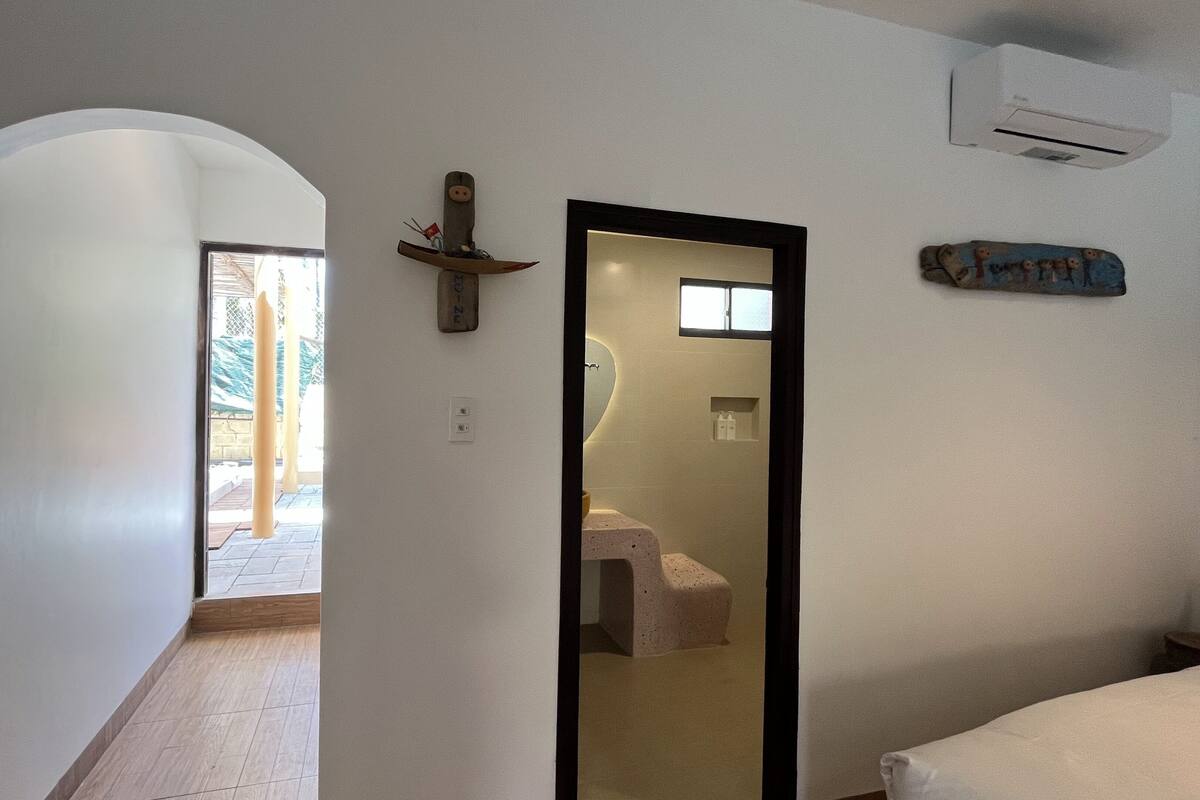 Um corredor estreito com um espelho, um banheiro pequeno com vaso sanitário e pia, e uma porta que dá para área externa com vista para um jardim.