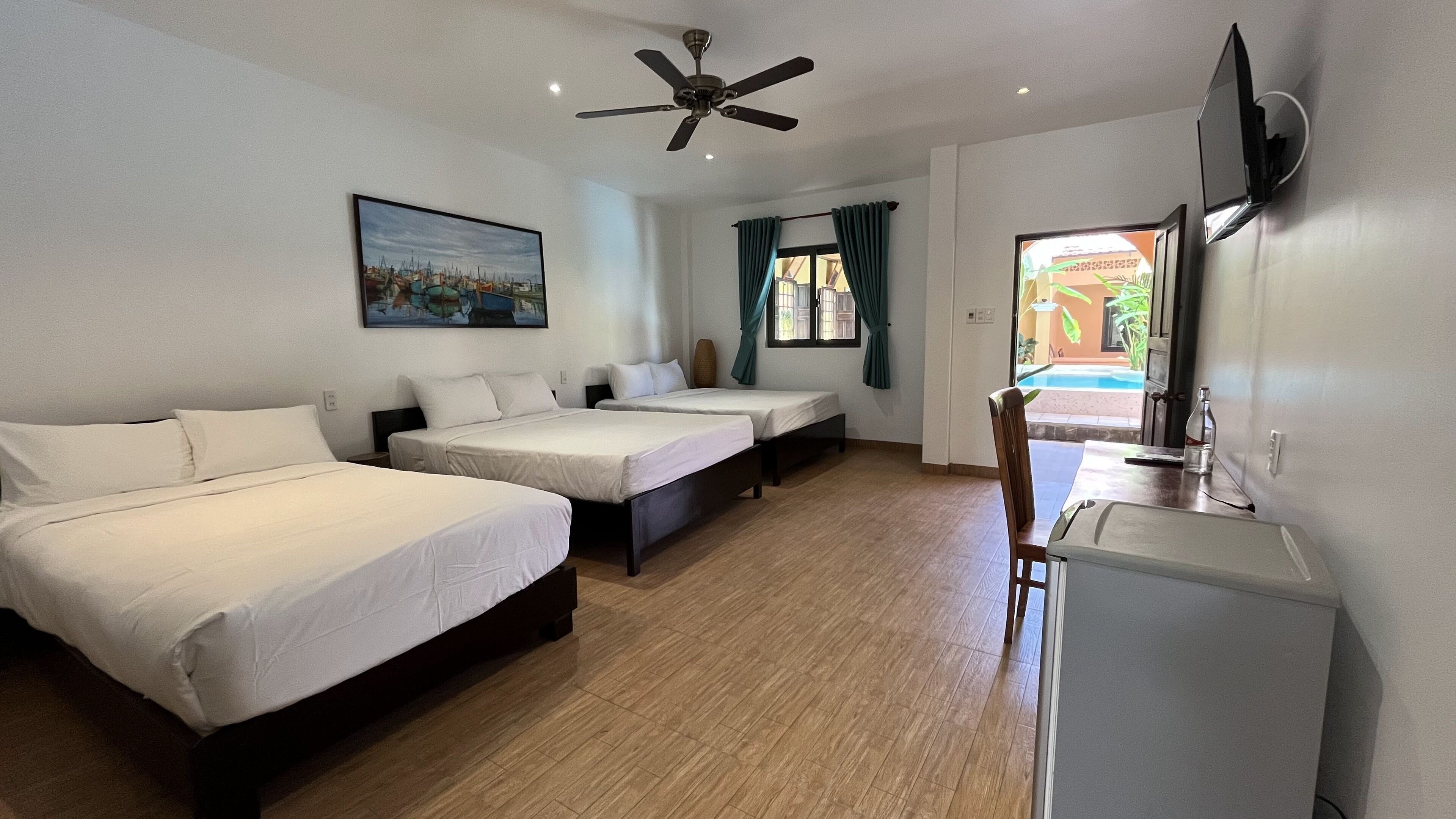 A hotel room with two single beds, a desk with a chair, a ceiling fan, a painting on the wall, and a window with curtains.