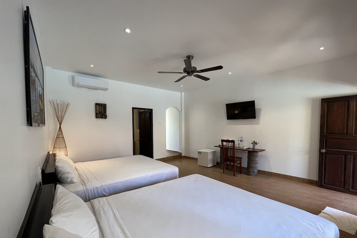 Quarto de hotel com duas camas, uma escrivaninha, uma cadeira, uma televisão e um ventilador de teto.