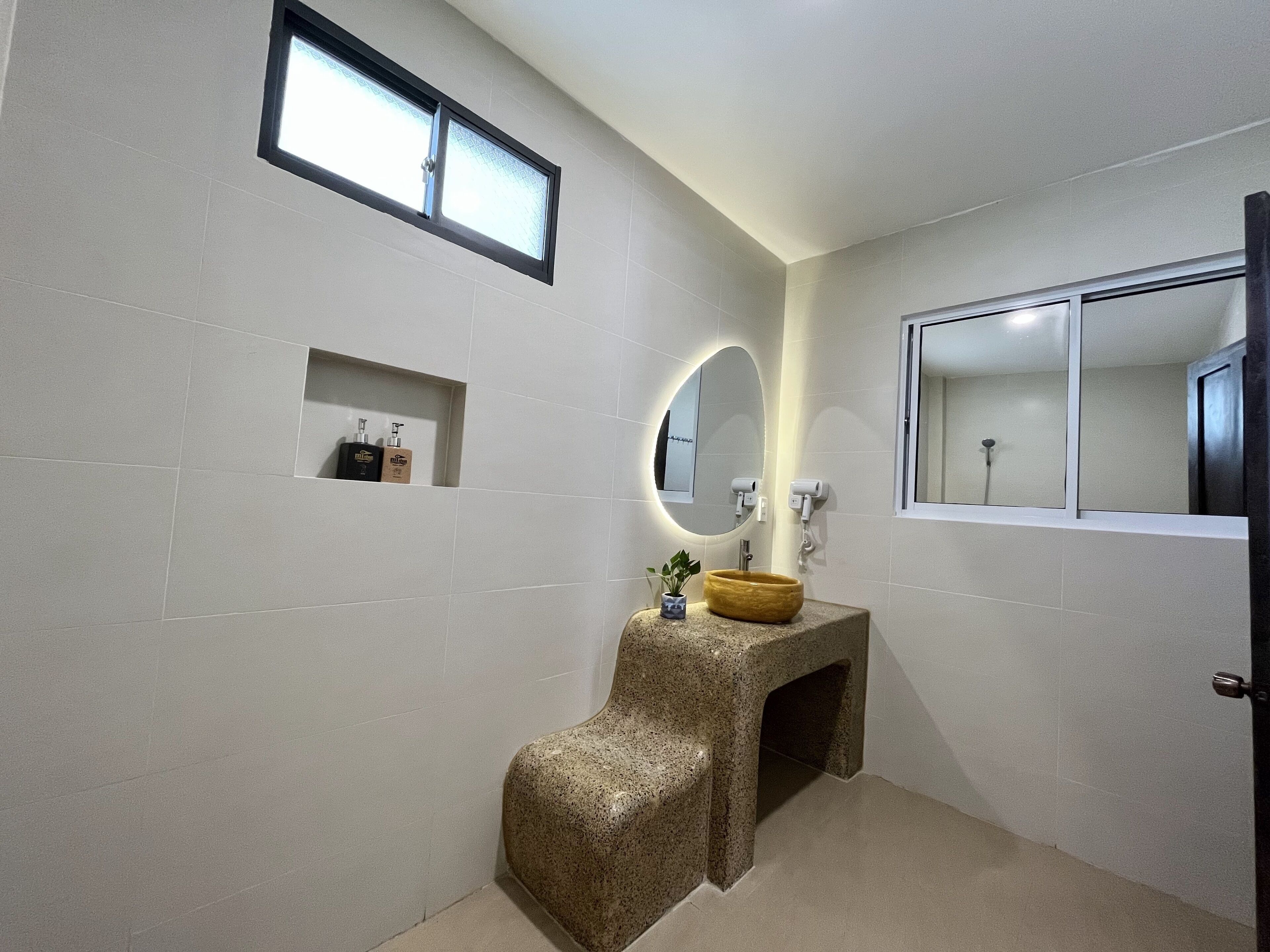 Une salle de bain avec un meuble-lavabo en granit, un miroir rond, une petite étagère contenant des articles de toilette et une fenêtre avec un panneau en verre dépoli.