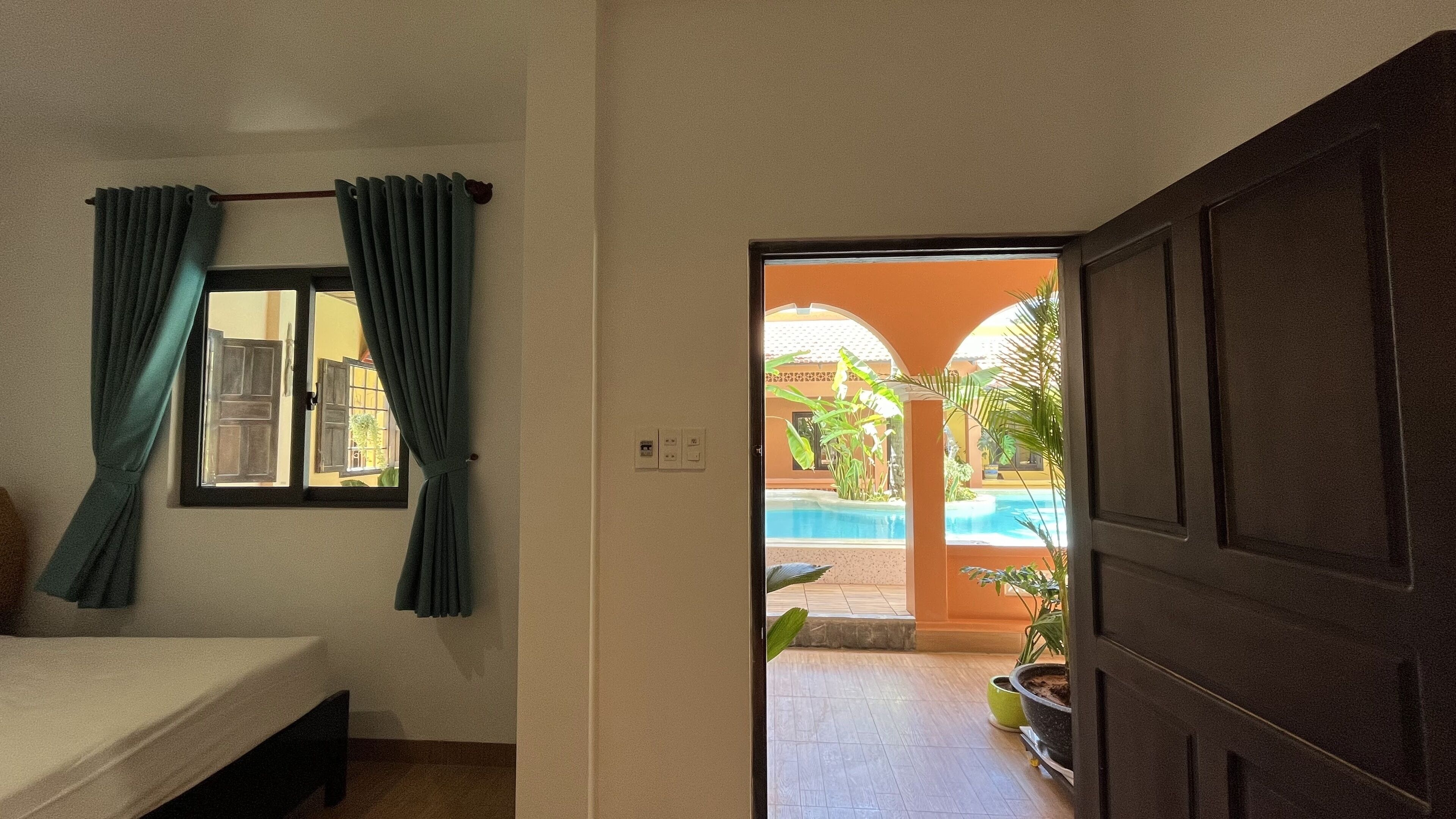 A room with a bed, a window with curtains, and an open door leading to an outdoor area with a pool and plants.