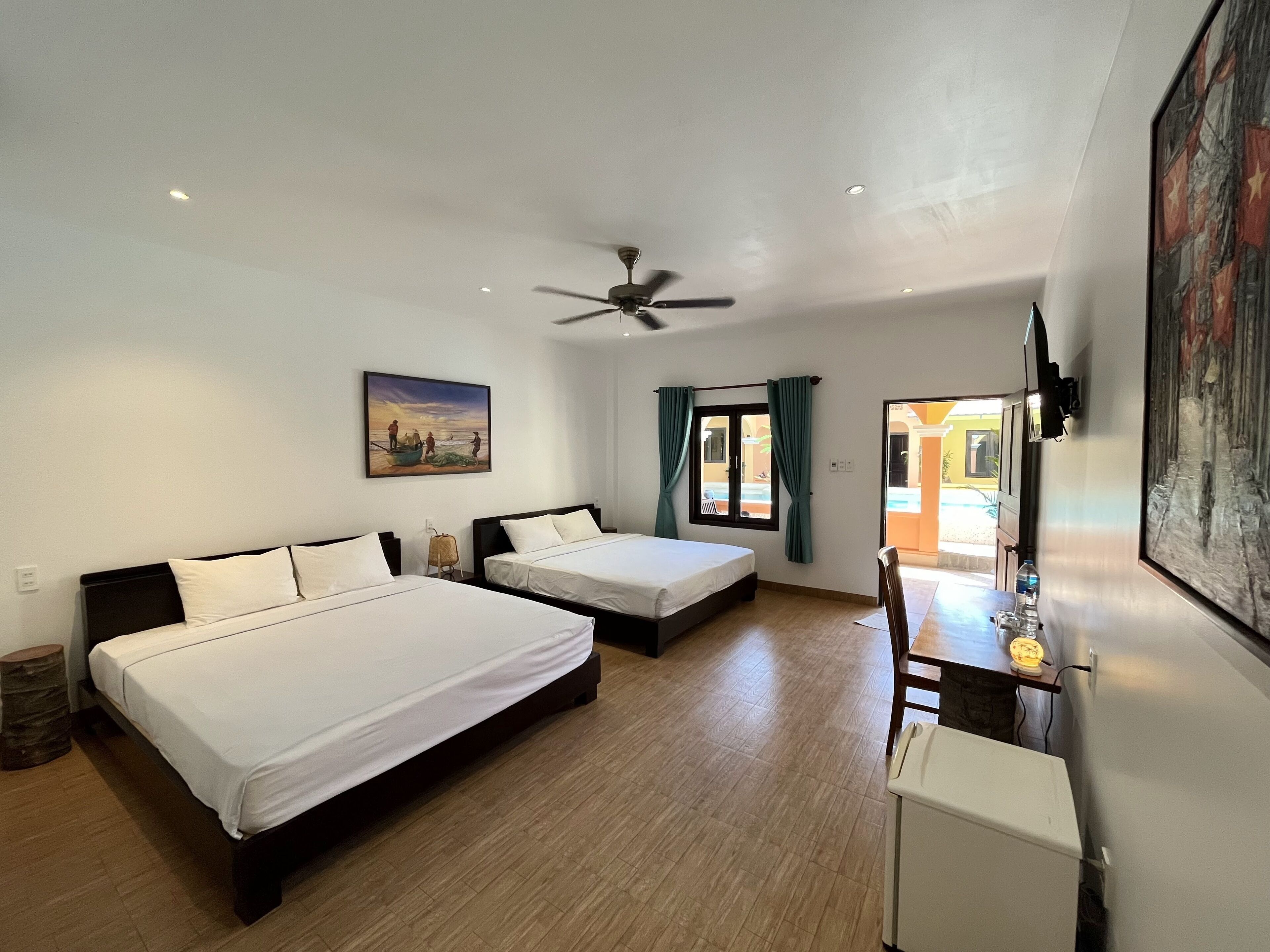 A hotel room with two beds, a ceiling fan, a painting, a window with curtains, and a view of the outside.