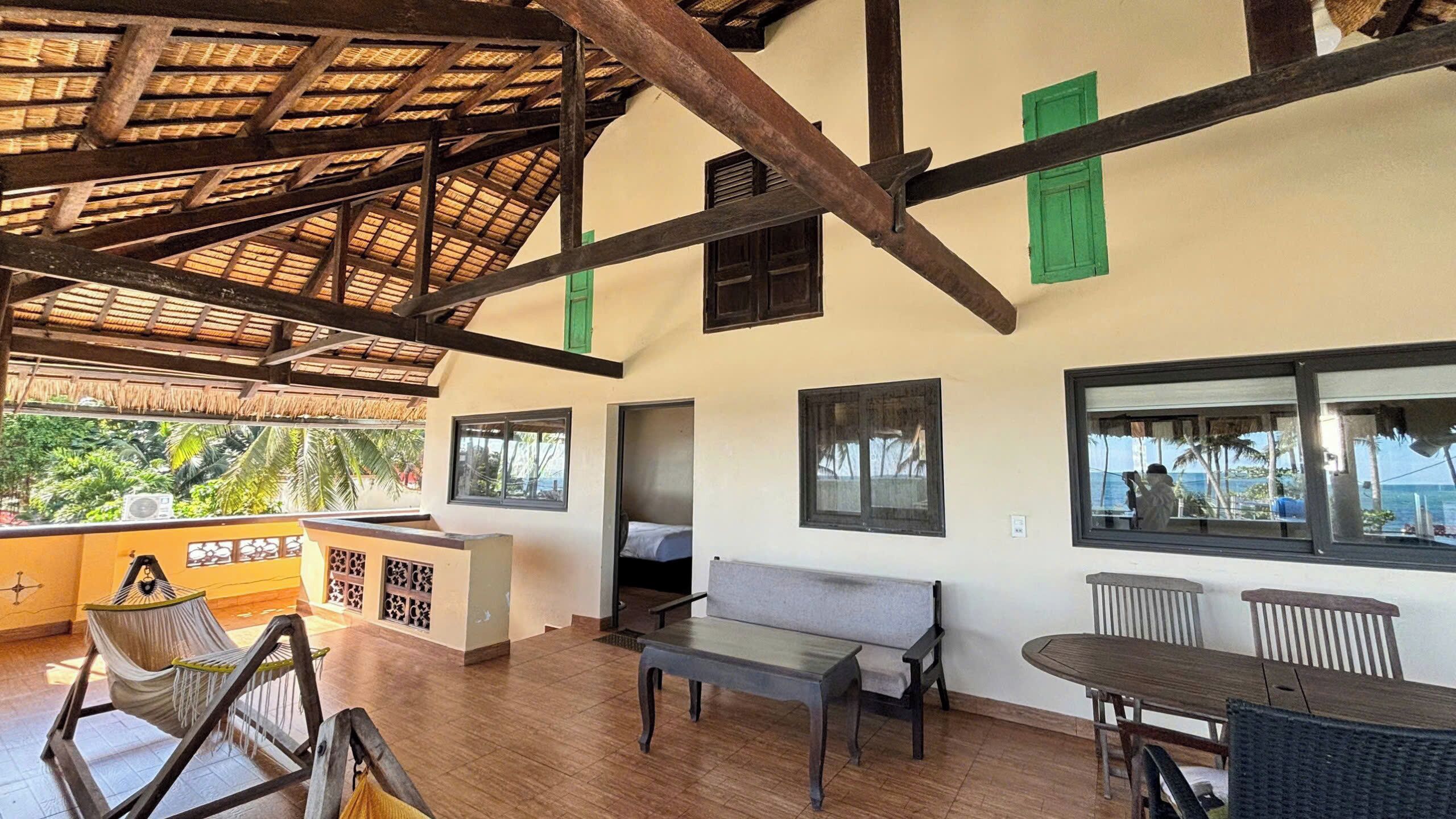 A spacious room with a high ceiling, wooden beams, a thatched roof, a sofa, a table, and a view of the outdoors.