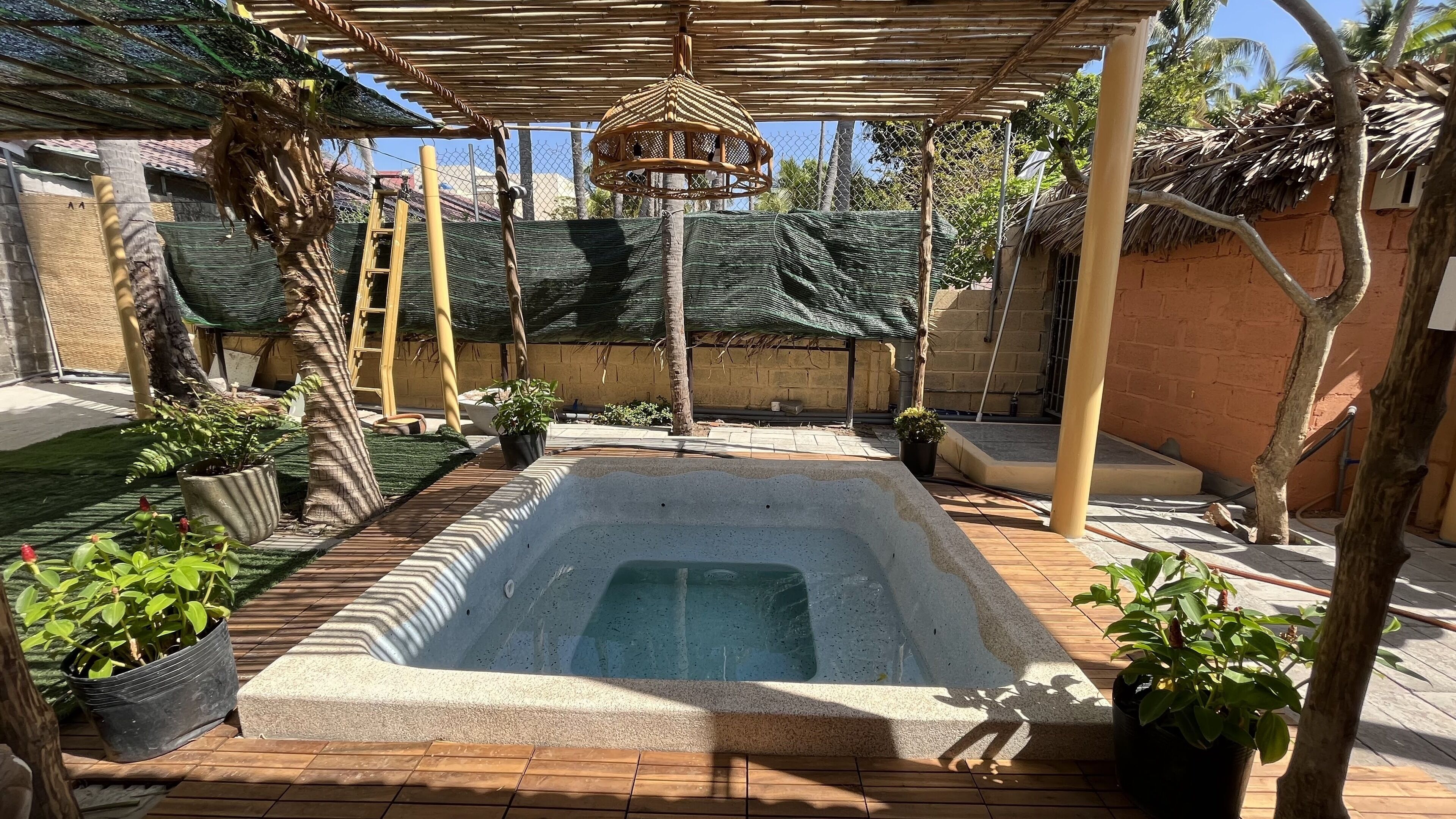 A rectangular hot tub with a wooden deck surrounding it, a pergola overhead, and potted plants.