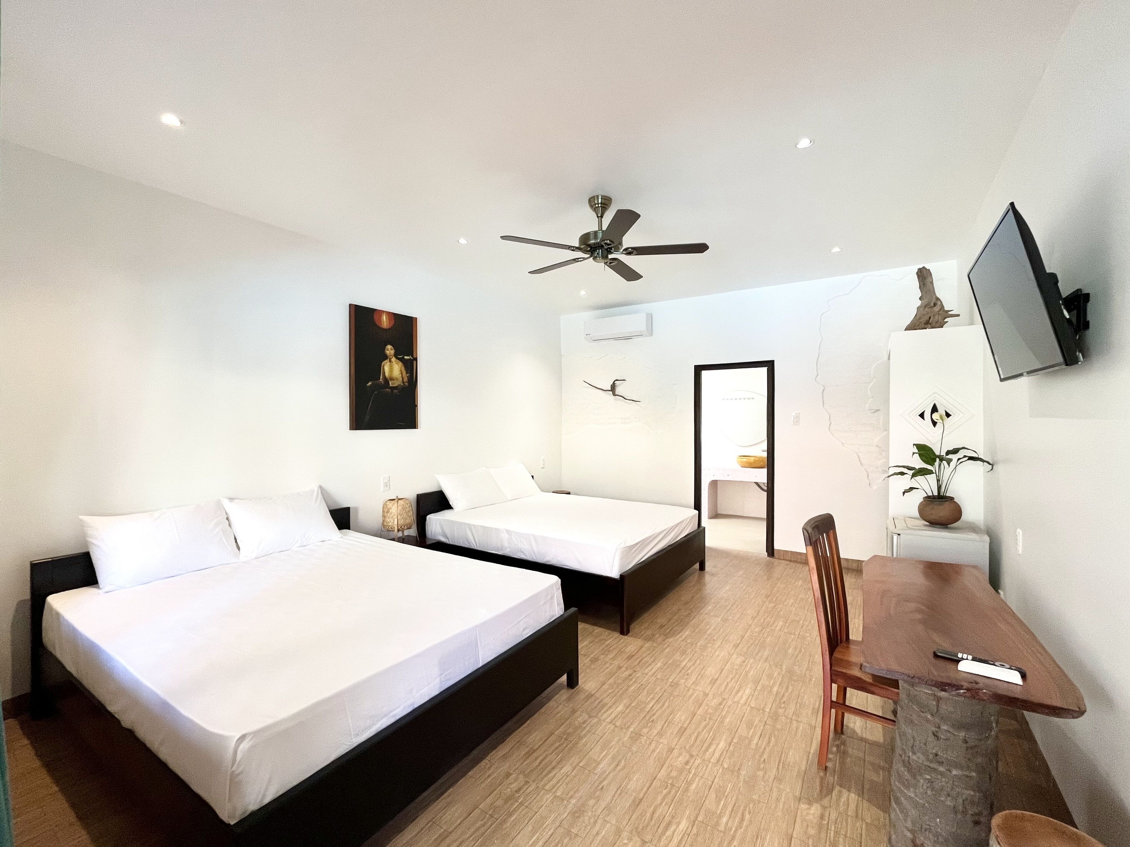 A hotel room with two beds, a desk, a chair, a ceiling fan, a TV, and a painting on the wall.