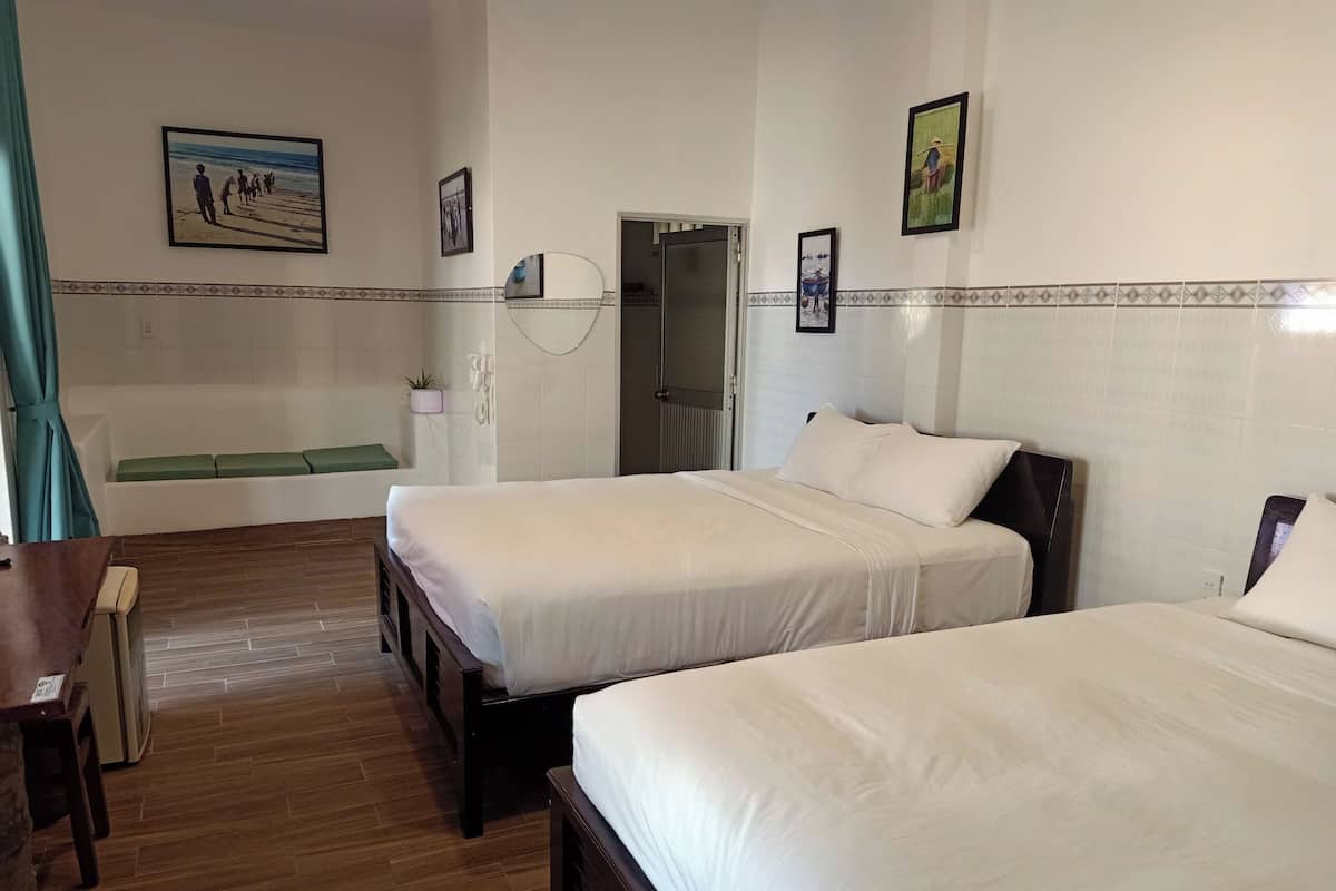 Quarto de hotel com duas camas, chão de madeira, um banco verde e parede com quadros emoldurados.