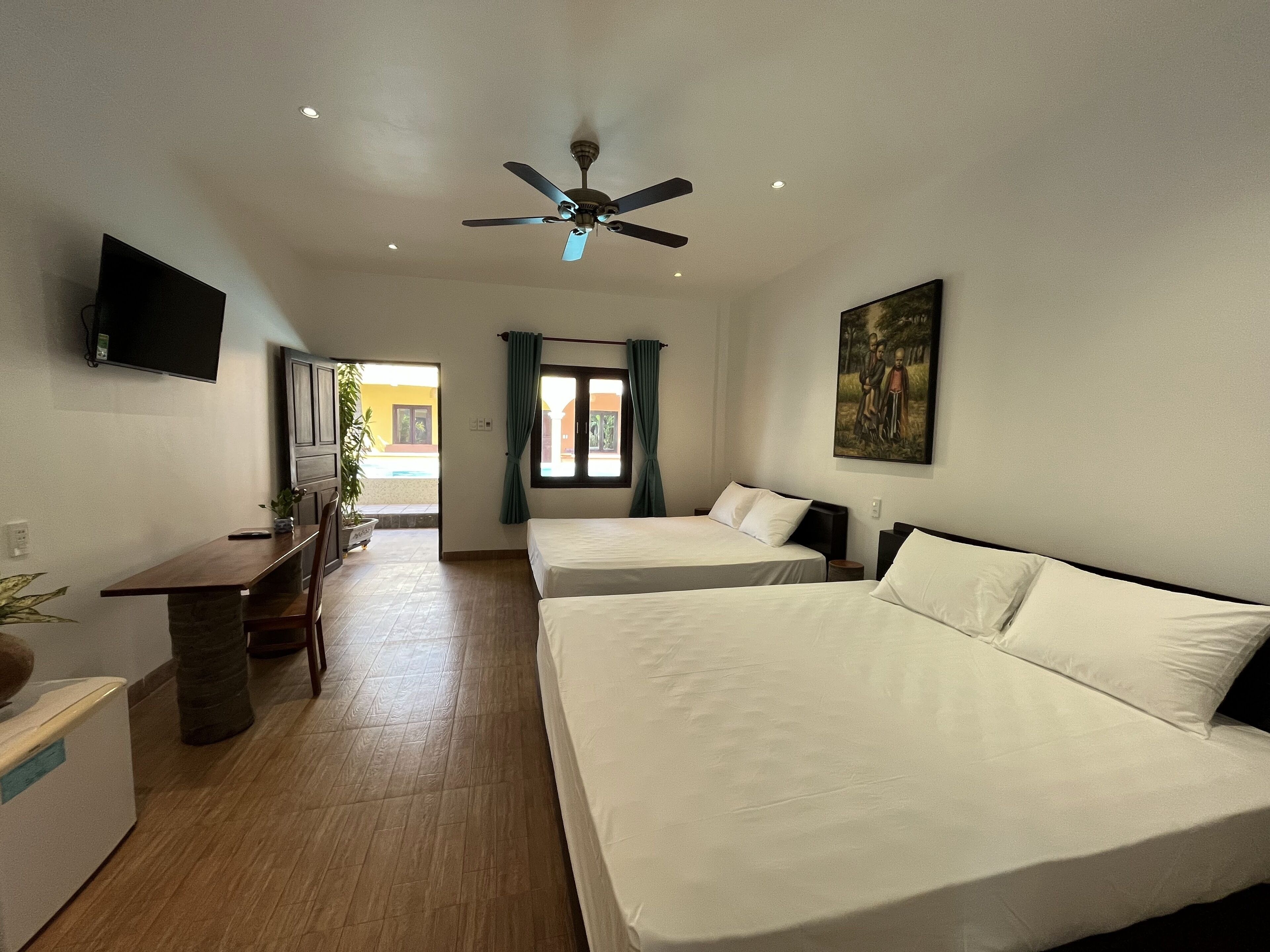 A hotel room with two beds, a desk, a ceiling fan, a TV, and a painting on the wall.