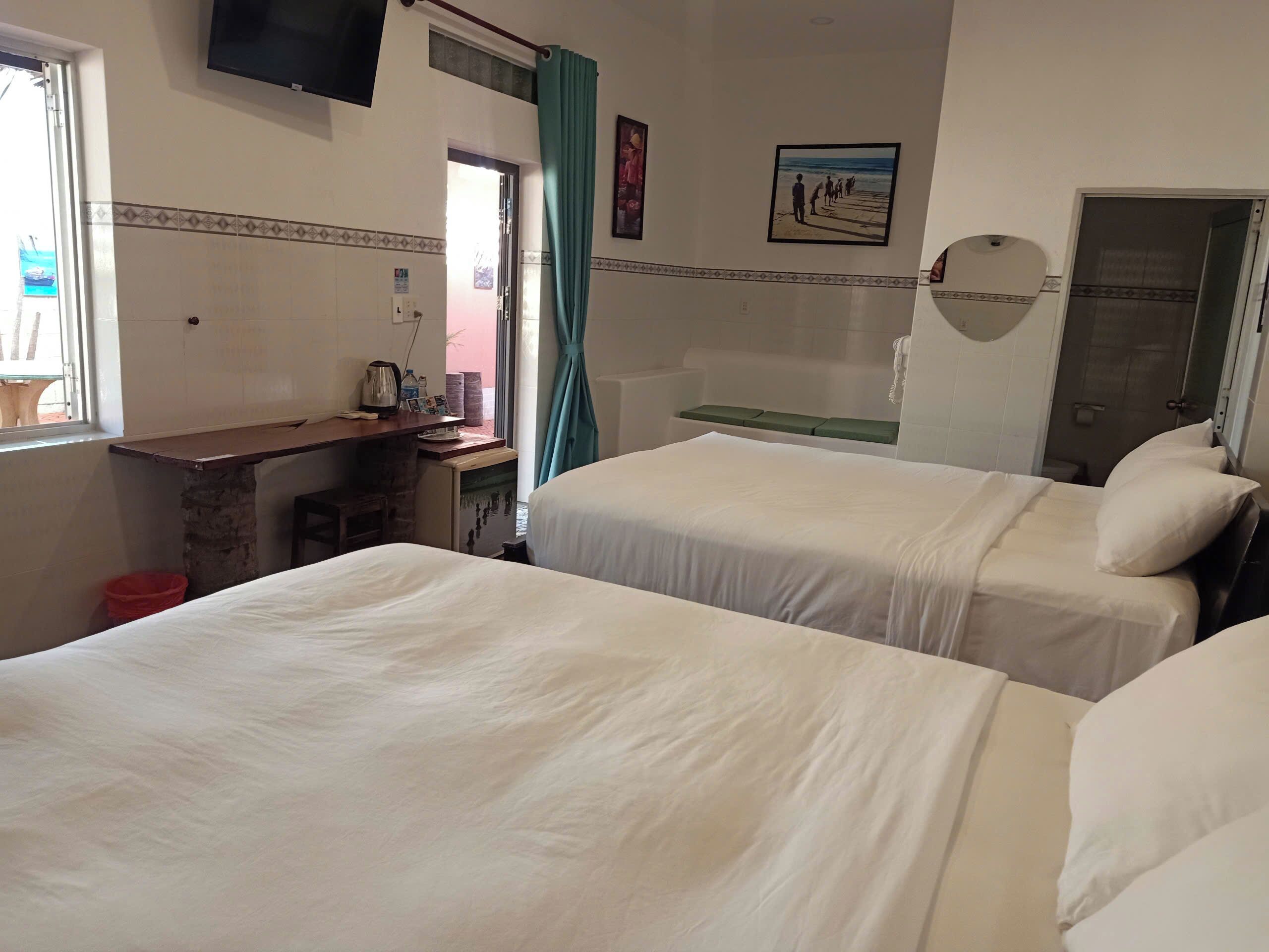 A hotel room with two beds, a desk with a chair, a small table, a TV mounted on the wall, and a shower area.