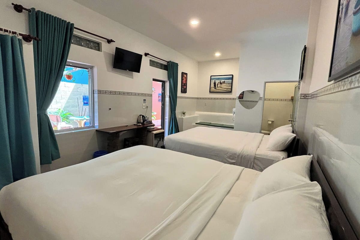 Quarto de hotel com duas camas, televisão, janela com cortinas, mesa pequena e banheiro com banheira e espelho.