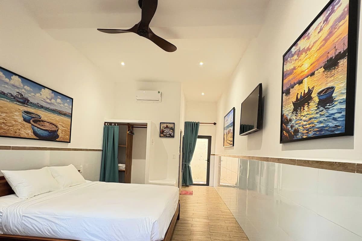 Quarto de hotel com uma cama grande, piso de madeira, ventilador de teto e obras de arte nas paredes.