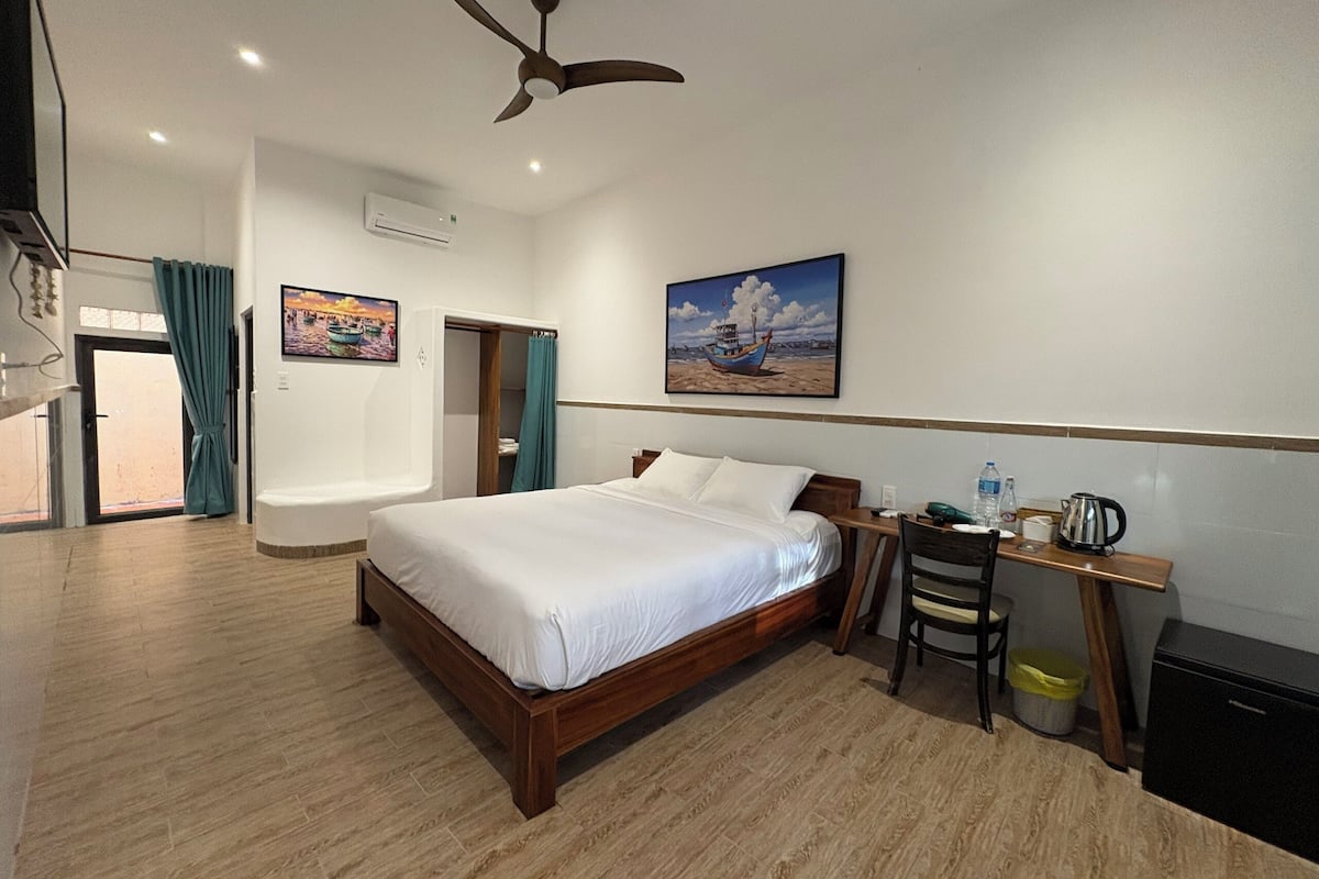 Quarto de hotel com cama, escrivaninha, cadeira, ventoinha no teto e um quadro na parede.